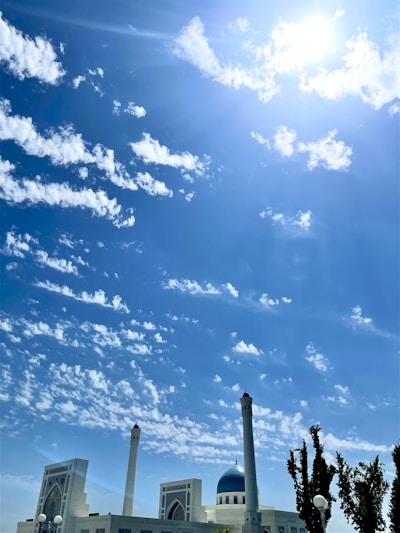A mosque with tall minarets and a blue dome stands prominently against a backdrop of a bright blue sky filled with scattered white clouds. The sun shines brightly, casting a warm glow over the scene. There are trees to the side, adding greenery to the foreground.