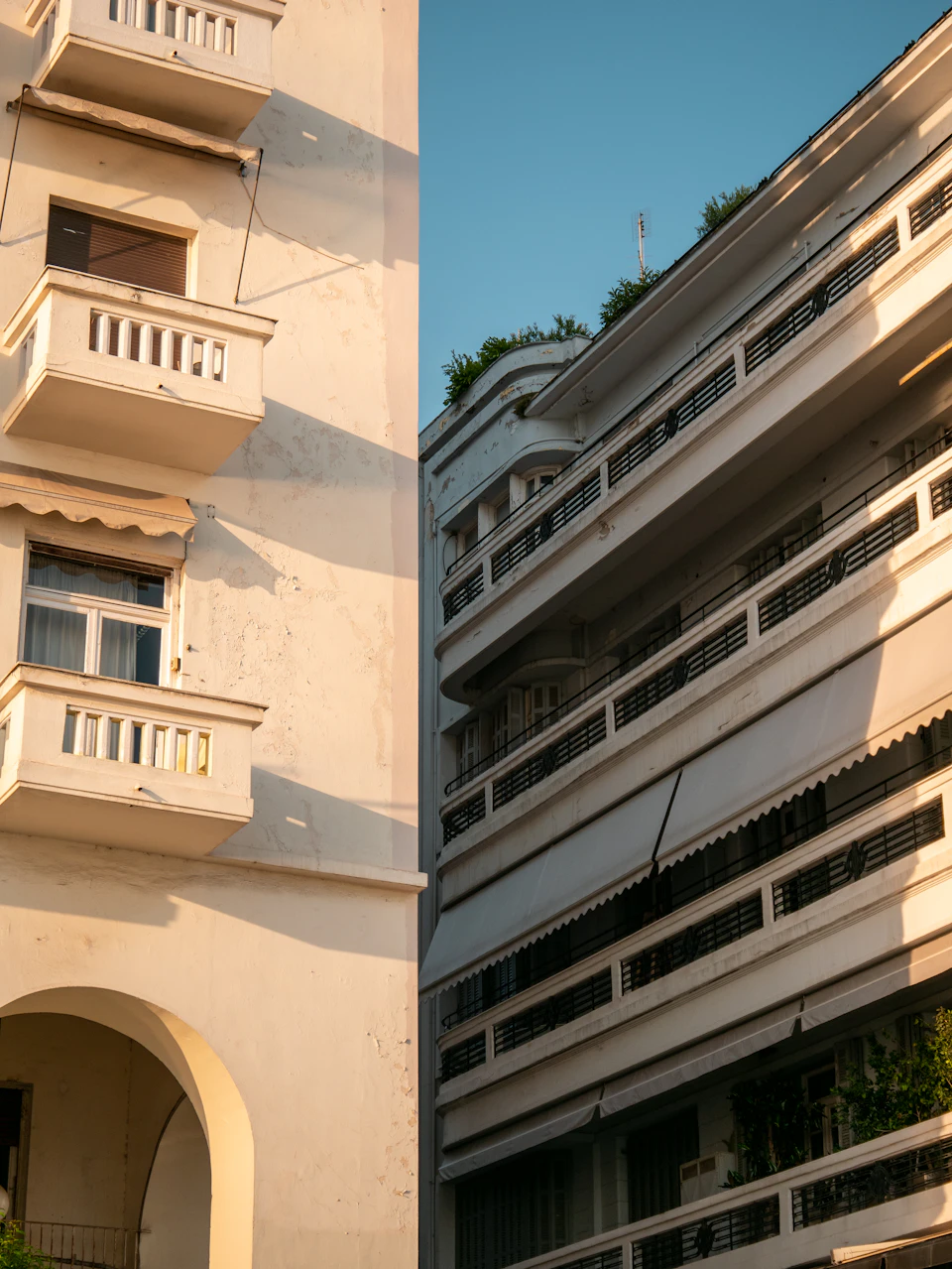 The image shows a two-story white building with three balconies on each floor, situated between another two-story white building and a tree. The first floor balcony is adorned with a white railing, while the second floor balcony has a black railing. The third floor balcony features a green plant.