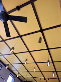 A ceiling features a grid-like pattern with large, alternating squares painted in yellow. There is a black ceiling fan with three blades, and multiple hanging pendant lights with exposed bulbs suspended at different lengths. The architecture presents a modern and stylish design.