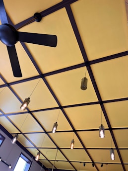A ceiling features a grid-like pattern with large, alternating squares painted in yellow. There is a black ceiling fan with three blades, and multiple hanging pendant lights with exposed bulbs suspended at different lengths. The architecture presents a modern and stylish design.
