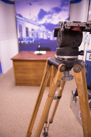 A wooden tripod is in the foreground with a camera mount on top, positioned in a room resembling a studio setup. In the background, there is a desk with a microphone on top and a chair behind it. The backdrop displays an image of a building with blue hues, possibly simulating an outdoor setting.