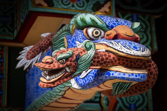 A detailed close-up of a vibrantly painted, traditional dragon sculpture with intricate carvings. The dragon features bold colors such as blue, red, and green, with large eyes and an expressive face that includes detailed scales and sharp teeth.