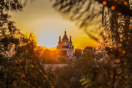 A picturesque church is silhouetted against a vibrant sunset, surrounded by lush trees, creating a serene atmosphere. The sun casts a golden glow, illuminating the sky with warm hues. The scene is framed by blurred foliage in the foreground, adding depth to the composition.
