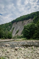 A tranquil landscape featuring a rocky riverbed with flowing water in the foreground. Tall, lush green trees cover the hillside on both sides, and a large cliff with exposed rock layers rises in the background under a partly cloudy sky.
