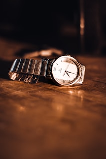 A close-up of a wristwatch with a metal strap lying on a wooden surface. The watch face has Roman numerals and a simple design, reflecting warm light that creates a vintage, elegant atmosphere.