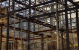 A skeletal structure of metal beams within a natural outdoor setting. The unfinished framework allows views of the surrounding trees and distant mountains, with beams creating geometric patterns. Sunlight filters through the gaps, casting shadows and highlighting parts of the structure.