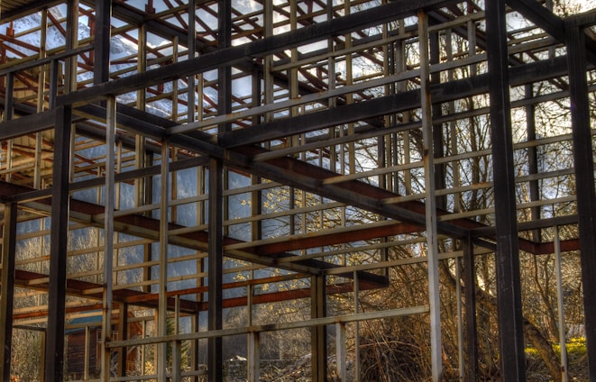 A skeletal structure of metal beams within a natural outdoor setting. The unfinished framework allows views of the surrounding trees and distant mountains, with beams creating geometric patterns. Sunlight filters through the gaps, casting shadows and highlighting parts of the structure.