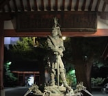 A detailed statue of a warrior in traditional armor stands prominently. The figure holds a calm yet commanding pose under an ornate roof, surrounded by greenery. The statue features intricate designs, including a dragon motif on the headgear. A dimly lit backdrop with trees and structural elements adds depth to the scene.