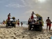 Several lifeguards in red shorts are standing around two off-road vehicles on a sandy beach. The vehicles are equipped with rescue equipment, including surfboards and flotation devices. In the background, people are enjoying the ocean, some swimming and others walking along the shore. The sky is partly cloudy with the sun breaking through.