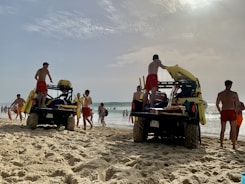 Several lifeguards in red shorts are standing around two off-road vehicles on a sandy beach. The vehicles are equipped with rescue equipment, including surfboards and flotation devices. In the background, people are enjoying the ocean, some swimming and others walking along the shore. The sky is partly cloudy with the sun breaking through.