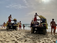 Several lifeguards in red shorts are standing around two off-road vehicles on a sandy beach. The vehicles are equipped with rescue equipment, including surfboards and flotation devices. In the background, people are enjoying the ocean, some swimming and others walking along the shore. The sky is partly cloudy with the sun breaking through.
