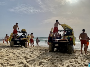 Several lifeguards in red shorts are standing around two off-road vehicles on a sandy beach. The vehicles are equipped with rescue equipment, including surfboards and flotation devices. In the background, people are enjoying the ocean, some swimming and others walking along the shore. The sky is partly cloudy with the sun breaking through.