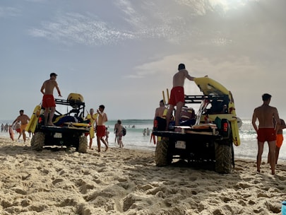 Several lifeguards in red shorts are standing around two off-road vehicles on a sandy beach. The vehicles are equipped with rescue equipment, including surfboards and flotation devices. In the background, people are enjoying the ocean, some swimming and others walking along the shore. The sky is partly cloudy with the sun breaking through.