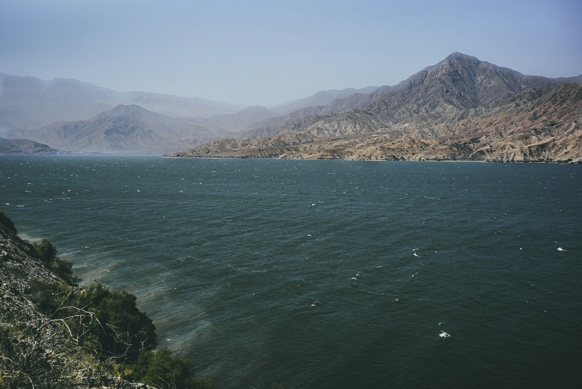 A calm, expansive body of water with gentle waves meets a rugged mountain landscape in the background. The shoreline is lined with scattered greenery, and the mountains display a mix of earthy tones with prominent ridges.