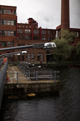 An industrial building with red brick architecture stands near the water. Several large pipes and a smokestack are visible, along with trees and greenery. The waterfront features a walkway with railings and several streetlights.
