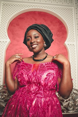 A woman is posing indoors, wearing an elegant, vibrant pink dress adorned with sequins and patterns. She dons a black head wrap and accessorizes with a black necklace and earrings. The background features a decorative wall with a white lattice archway and a pink backdrop.