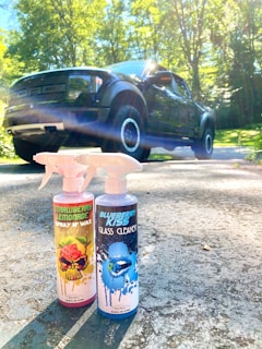Two bottles of car cleaning products labeled 'Strawberry Lemonade Spray N Wax' and 'Blueberry Kiss Glass Cleaner' are placed on a sunlit driveway with a large black pickup truck in the background. The scene is surrounded by green trees under a bright, clear sky.