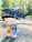 Two bottles of car cleaning products labeled 'Strawberry Lemonade Spray N Wax' and 'Blueberry Kiss Glass Cleaner' are placed on a sunlit driveway with a large black pickup truck in the background. The scene is surrounded by green trees under a bright, clear sky.