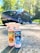 Two bottles of car cleaning products labeled 'Strawberry Lemonade Spray N Wax' and 'Blueberry Kiss Glass Cleaner' are placed on a sunlit driveway with a large black pickup truck in the background. The scene is surrounded by green trees under a bright, clear sky.
