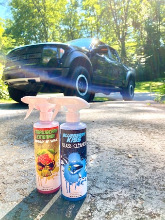 Two bottles of car cleaning products labeled 'Strawberry Lemonade Spray N Wax' and 'Blueberry Kiss Glass Cleaner' are placed on a sunlit driveway with a large black pickup truck in the background. The scene is surrounded by green trees under a bright, clear sky.