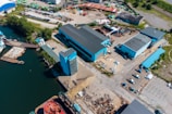 Aerial view of an industrial area featuring several blue-roofed buildings near a body of water. The complex includes large warehouses, an open yard with scattered machinery and vehicles, and loading docks. Adjacent to the structures are areas with greenery and paved roads, with a few parked cars and trucks scattered throughout. The waterway contains a pier and some small vessels.