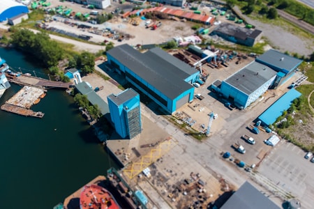 Aerial view of an industrial area featuring several blue-roofed buildings near a body of water. The complex includes large warehouses, an open yard with scattered machinery and vehicles, and loading docks. Adjacent to the structures are areas with greenery and paved roads, with a few parked cars and trucks scattered throughout. The waterway contains a pier and some small vessels.