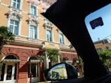 A grand historic hotel features ornate architectural details with decorative stucco and white highlights on a warm peach facade. Tall windows with elaborate moldings are present, and palm trees flank the entrance, creating a mix of elegance and tropical charm. A sign reads 'Mileni Hotel Continental'. The image is partially obscured by the interior of a car, as seen by the black silhouette and side view mirror reflecting parts of the hotel.