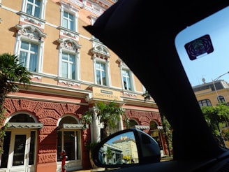 A grand historic hotel features ornate architectural details with decorative stucco and white highlights on a warm peach facade. Tall windows with elaborate moldings are present, and palm trees flank the entrance, creating a mix of elegance and tropical charm. A sign reads 'Mileni Hotel Continental'. The image is partially obscured by the interior of a car, as seen by the black silhouette and side view mirror reflecting parts of the hotel.