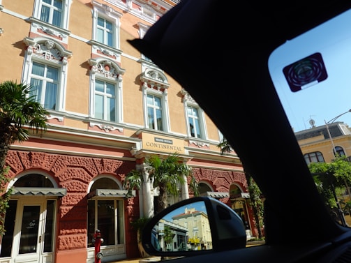 A grand historic hotel features ornate architectural details with decorative stucco and white highlights on a warm peach facade. Tall windows with elaborate moldings are present, and palm trees flank the entrance, creating a mix of elegance and tropical charm. A sign reads 'Mileni Hotel Continental'. The image is partially obscured by the interior of a car, as seen by the black silhouette and side view mirror reflecting parts of the hotel.