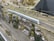 A model train runs along a detailed railway setup, featuring a sleek, silver train moving on tracks beside a rocky, grassy landscape. The foreground includes power lines and multiple tracks, while the background shows miniature trees and terrain.