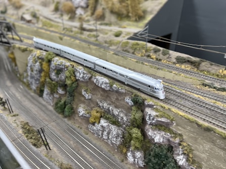 A model train runs along a detailed railway setup, featuring a sleek, silver train moving on tracks beside a rocky, grassy landscape. The foreground includes power lines and multiple tracks, while the background shows miniature trees and terrain.