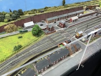 A detailed model railway scene features multiple train tracks running parallel with miniature train cars and a railway station. The setting includes model buildings resembling houses and industrial facilities, along with green landscaped areas, tree replicas, roads, and pathways. A backdrop of a painted blue sky and distant greenery completes the scene.