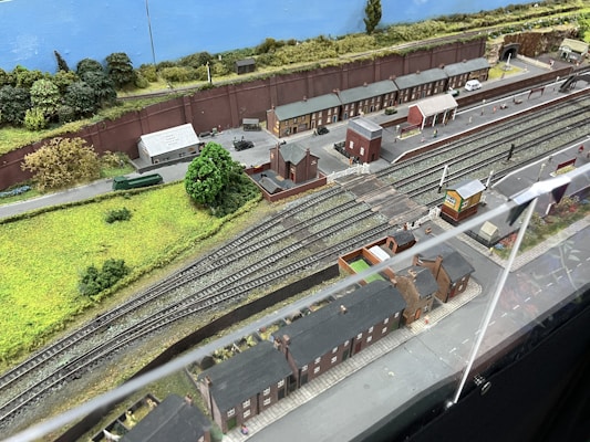 A detailed model railway scene features multiple train tracks running parallel with miniature train cars and a railway station. The setting includes model buildings resembling houses and industrial facilities, along with green landscaped areas, tree replicas, roads, and pathways. A backdrop of a painted blue sky and distant greenery completes the scene.