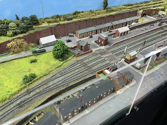A detailed model railway scene features multiple train tracks running parallel with miniature train cars and a railway station. The setting includes model buildings resembling houses and industrial facilities, along with green landscaped areas, tree replicas, roads, and pathways. A backdrop of a painted blue sky and distant greenery completes the scene.