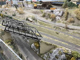 A detailed model train layout featuring a steel truss bridge over multiple railway tracks. The scene is set in a mountainous landscape with realistic terrain, including rocky hillsides and scattered vegetation. There are structures such as a wooden building and a warehouse in the background, along with model vehicles and utility poles. The layout captures the essence of a rural or industrial railway setting.