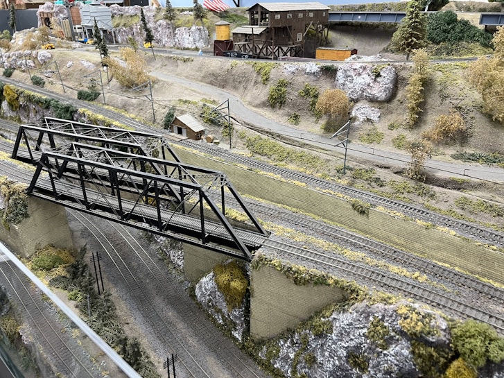A detailed model train layout featuring a steel truss bridge over multiple railway tracks. The scene is set in a mountainous landscape with realistic terrain, including rocky hillsides and scattered vegetation. There are structures such as a wooden building and a warehouse in the background, along with model vehicles and utility poles. The layout captures the essence of a rural or industrial railway setting.