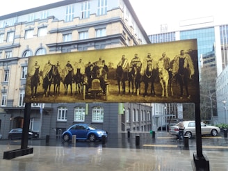 A large sepia-toned photograph in an outdoor setting features a group of men on horseback and an old car, displayed as an art installation. The photograph is set against an urban backdrop with a mixture of modern glass buildings and older architecture. Wet pavement suggests recent rain, and several modern cars are parked nearby.