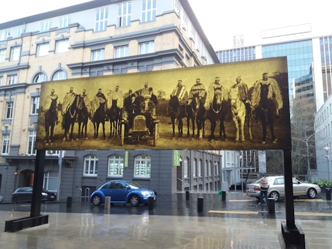 A large sepia-toned photograph in an outdoor setting features a group of men on horseback and an old car, displayed as an art installation. The photograph is set against an urban backdrop with a mixture of modern glass buildings and older architecture. Wet pavement suggests recent rain, and several modern cars are parked nearby.