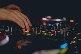 Close-up of a DJ's hands adjusting neon-lit controls on a mixing console in a dark studio.