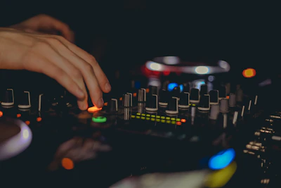 Close-up of a DJ's hands adjusting neon-lit controls on a mixing console in a dark studio.