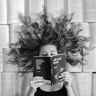 A woman lies surrounded by open books with her hair spread out in waves across the pages. She holds a book titled 'milk and honey' in front of her face, partially covering it. The image is in black and white, emphasizing the contrast between the text and her wavy hair.