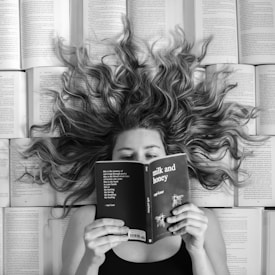 A woman lies surrounded by open books with her hair spread out in waves across the pages. She holds a book titled 'milk and honey' in front of her face, partially covering it. The image is in black and white, emphasizing the contrast between the text and her wavy hair.