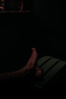 A foot is illuminated in a dimly lit environment, with shadows cast across the floor or surface. The scene is dark, with a deep contrast between the light and shadowed areas.