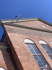 A red brick building with large windows and an intricate cornice under a clear blue sky. Security cameras are mounted at the top corner.