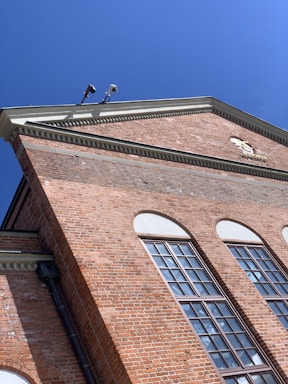 A red brick building with large windows and an intricate cornice under a clear blue sky. Security cameras are mounted at the top corner.