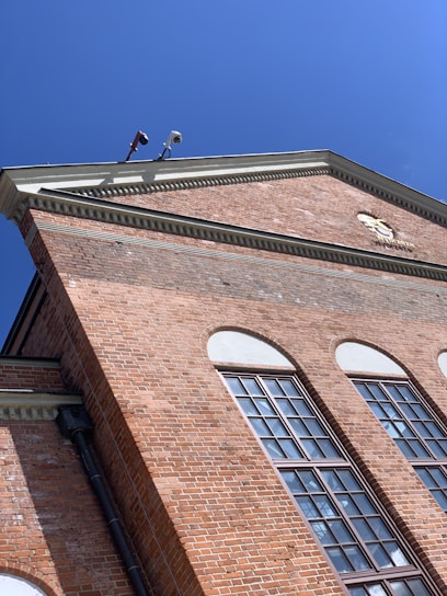 A red brick building with large windows and an intricate cornice under a clear blue sky. Security cameras are mounted at the top corner.