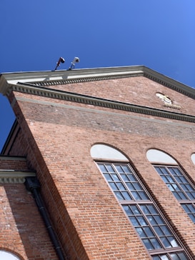 A red brick building with large windows and an intricate cornice under a clear blue sky. Security cameras are mounted at the top corner.