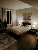 A modern, cozy bedroom with a wooden bed frame adorned with white bedding and pillows. Two bedside tables with lamps flank the bed, casting a warm light. Above the bed hang two prints featuring deer. A large window with drawn curtains is on the left, and a patterned rug lies on the wooden floor.