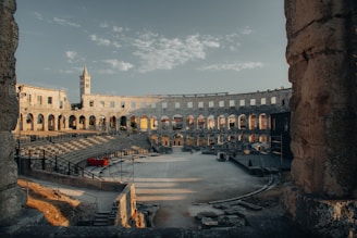 An ancient Roman amphitheater with large stone arches and a partially intact exterior wall is seen under a blue sky with white clouds. The interior shows tiered stone seating surrounding a central arena space. A tower is visible in the background, partially illuminated by the setting sun.