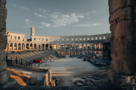 An ancient Roman amphitheater with large stone arches and a partially intact exterior wall is seen under a blue sky with white clouds. The interior shows tiered stone seating surrounding a central arena space. A tower is visible in the background, partially illuminated by the setting sun.